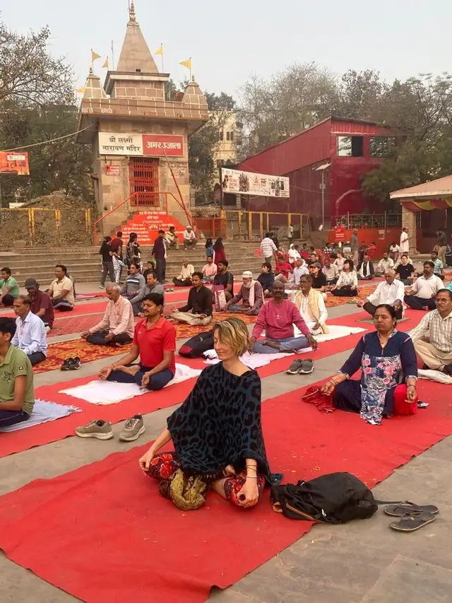 Marie-Laure assise en méditation sur un tapis rouge au bord du Gange, devant des pratiquants indiens